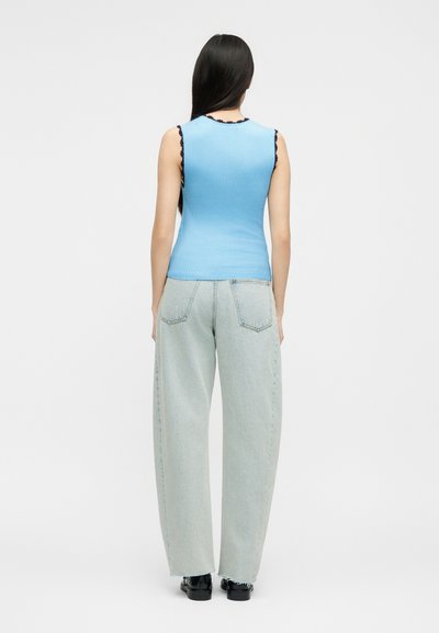 Γυναίκα με μακριά σκούρα μαλλιά που φορά αμάνικη μπλε ριμπ τοπ και ανοιχτόχρωμα τζιν με φαρδιά πόδια, στέκεται πλάτη στον θεατή.