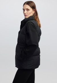 Eine Frau in einer schwarzen, gesteppte ärmellosen Weste und einem langärmligen Oberteil steht seitlich mit den Händen in den Taschen vor einem einfarbigen grauen Hintergrund.