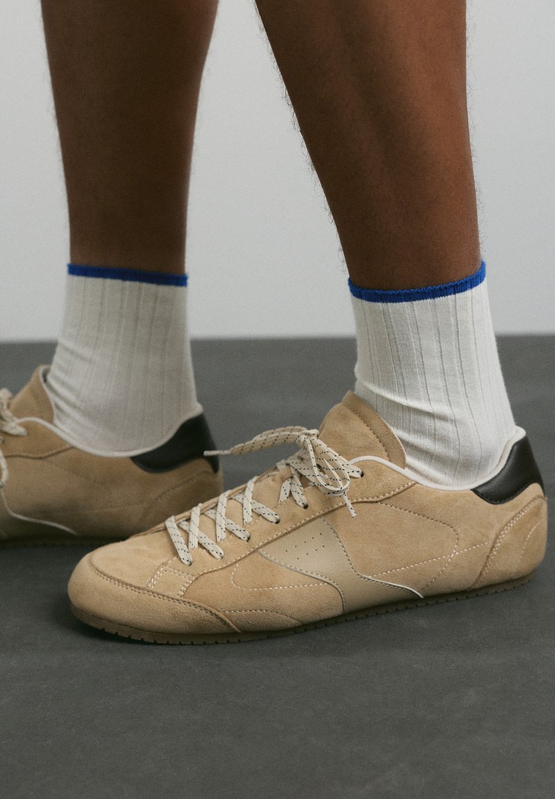 Jambes portant des chaussettes blanches côtelées avec un bord bleu et des baskets en suède beige avec des accents noirs au talon et des lacets blancs sur un sol gris.