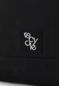 Black fabric label with "recycle" in white text, featuring a textured background and stitched edges, attached to a black canvas item.