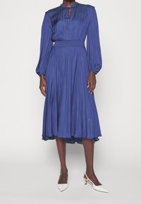 Vestido plisado de un azul profundo con mangas largas, cuello atado y cintura elástica; combinado con tacones blancos de punta. 