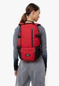 Donna con coda di cavallo che indossa uno zaino Eastpak rosso con spallacci e cerniere nere, vestita con una maglietta a maniche lunghe grigio e nera sfumata e pantaloni grigi.