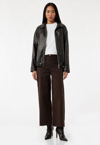 Chaqueta de cuero sintético negra con cuello, camisa blanca y pantalones de pierna ancha marrones. Acentuada con una cremallera y tacones. Textura suave.