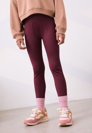 Persona che indossa leggings a coste color granata, calzini rosa chiaro, sneakers rosa e beige e una felpa beige corta, in piedi su un pavimento grigio.