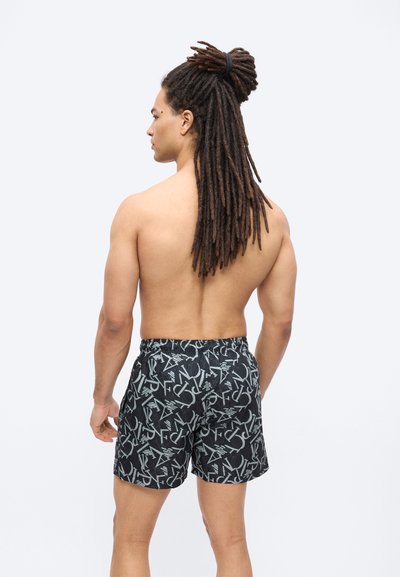 Άνδρας χωρίς πουκάμισο με μακριά κοτσίδα δεμένα ψηλά, φοράει μαύρο σορτς με μεγάλο, αλληλοεπικαλυπτόμενο μοτίβο ανοιχτόγριων γραμμάτων, γυρισμένος προς τα πίσω.