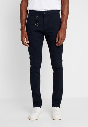 Hombre con pantalones ajustados azul marino oscuro con cadena de llavero, zapatillas blancas y camisa blanca metida por dentro, de pie contra un fondo liso.