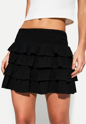 Woman wearing a black tiered ruffle mini skirt with a smocked waistband and a white cropped top, showing midriff and legs.