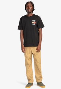 Zwarte katoenen t-shirt met een ontwerp van een hart en slot, gecombineerd met beige cargobroek en grijze sneakers met gele zolen.