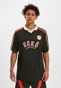 Tricou negru cu un guler alb în contrast, accente portocalii pe mâneci și logo-ul "ECKŌ unlimited" pe față. Textură de material moale.