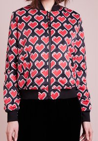 Black bomber jacket featuring red pixelated heart patterns, zip closure, ribbed cuffs, and waistband in a glossy, smooth fabric.
