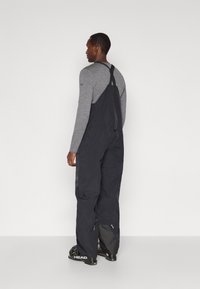 Άνδρας με μαύρες φόρμες σκι και γκρι μπλούζα με μακρύ μανίκι, εικονιζόμενος από πίσω, να στέκεται σε λευκό φόντο.