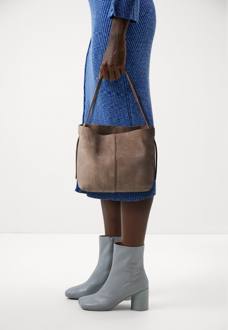 Personne portant une robe côtelée bleue et des bottines gris clair tenant un sac à main en daim marron par ses poignées à leur côté.