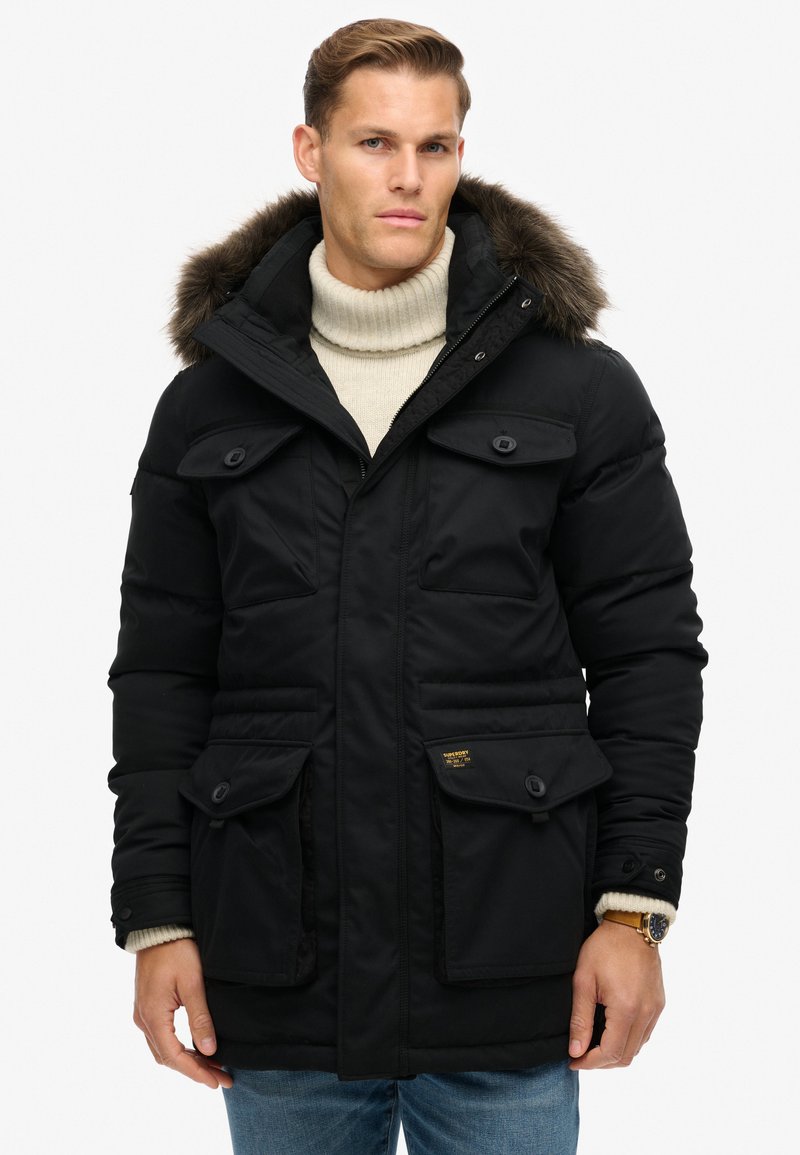 Zwarte parka-jack met een met bont gevoerde capuchon, een rits aan de voorkant en knoopjes op de borstzakken. Heeft een gladde, geïsoleerde textuur en verstelbare manchetten.