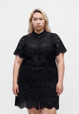 Femme aux longs cheveux blonds portant une robe noire en dentelle œillet à manches courtes avec une patte de boutonnage devant, debout devant un fond blanc uni.