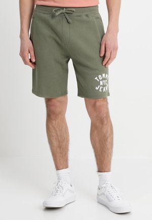 Parte inferiore del corpo di una persona che indossa pantaloncini verde oliva con coulisse e testo bianco "Tommy NYC Jeans" e scarpe da ginnastica bianche con calzini su sfondo uniforme.