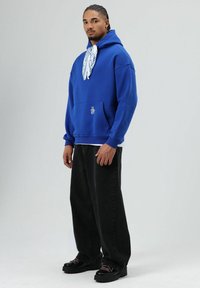 Blauwe hoodie met trekkoord en voorzak, gecombineerd met losse zwarte broek en stevige zwarte schoenen. Witte sjaal met patroon om de nek gebonden.