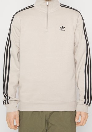 Béžová mikina Adidas s čtvrtkruhovým zipem, s černými třemi pruhy na rukávech a malým černým logem Adidas na hrudi.