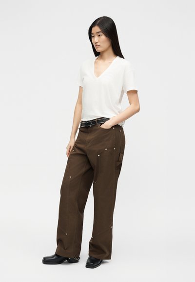 Γυναίκα που στέκεται με το χέρι στην τσέπη, φοράει λευκό κοντομάνικο μπλουζάκι με V λαιμόκοψη, καφέ παντελόνι φαρδύ στο πόδι, μαύρη ζώνη και μαύρα παπούτσια.