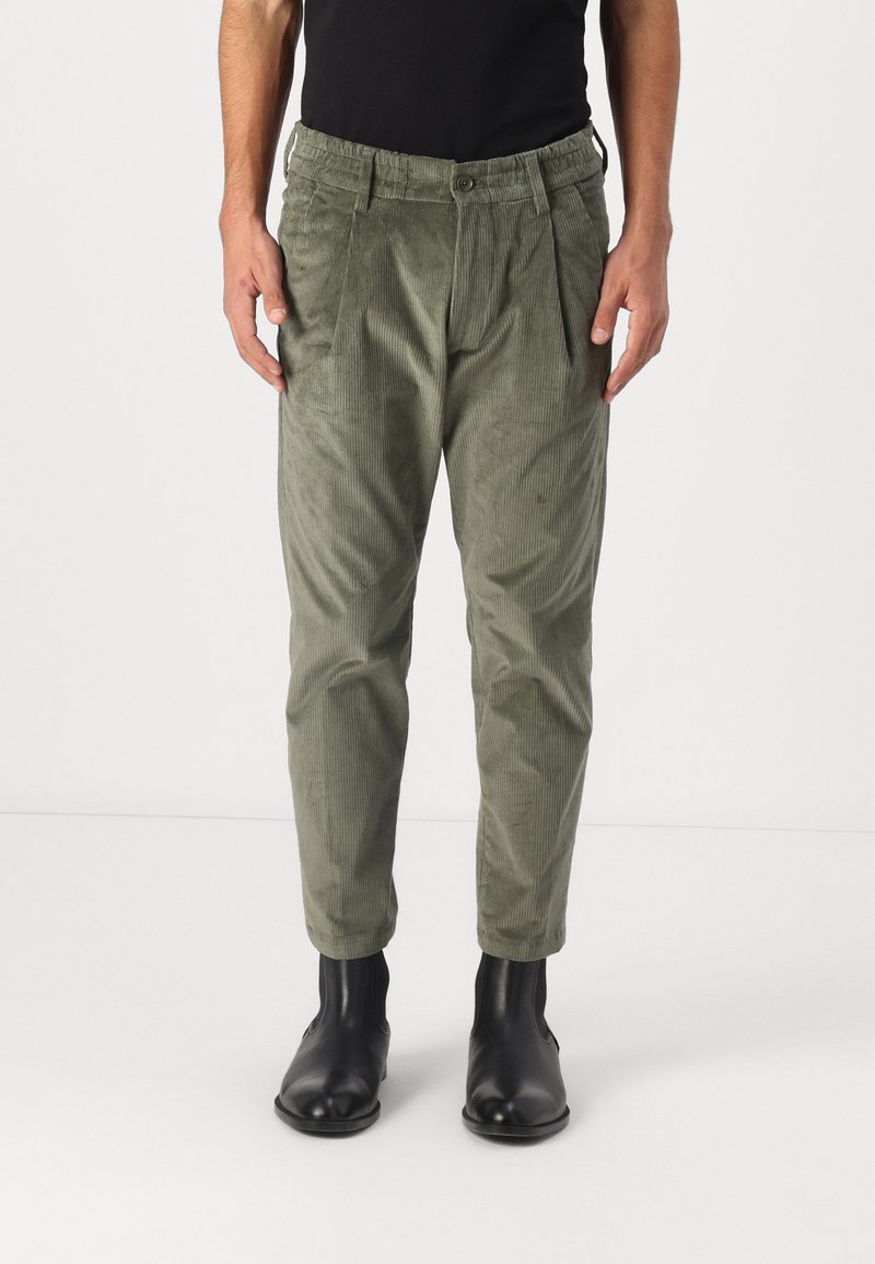 Pantalons en velours côtelé vert olive avec une coupe décontractée, dotés d'une taille à bouton et élastique, de plis et de jambes fuselées, associés à des bottines noires.