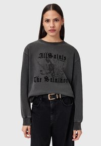 Donna con capelli lisci e scuri che indossa una felpa nera sbiadita con uno scheletro e il testo "AllSaints The Sainthood", insieme a jeans neri con cintura.