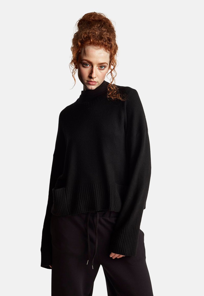 Schwarzer Oversize-Pullover mit geripptem Saum und Bündchen, ausgestattet mit einem hohen Kragen und einer lockeren Passform. Sanfte Textur, kombiniert mit dunklen Hosen.