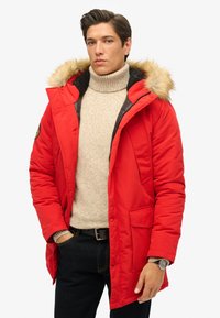Czerwona parka z futrzanym wykończeniem kaptura, zapinana na zamek błyskawiczny z dwoma przednimi kieszeniami. W komplecie z kremowym golfem i ciemnymi dżinsami.