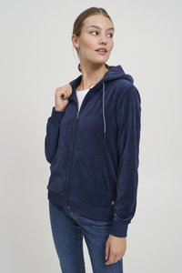 Tmavomodrá mikina na zips s kapucňou, bočnými vreckami a sťahovacou šnúrkou, vyrobená z mäkkej textúry. Kombinovaná s modrými rifľovými nohavicami.