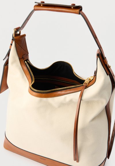 Κρεμ τσάντα ώμου από καμβά με καφέ δερμάτινες λεπτομέρειες, χρυστό φερμουάρ και ρυθμιζόμενο λουρί, εμφανιζόμενη μερικώς ανοιχτή και ορατή μαύρη εσωτερική επένδυση.