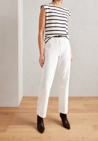 Pantalones blancos ajustados con pierna recta y bolsillos laterales, combinados con una blusa sin mangas de rayas blancas y negras. Incluidos botines negros de punta.