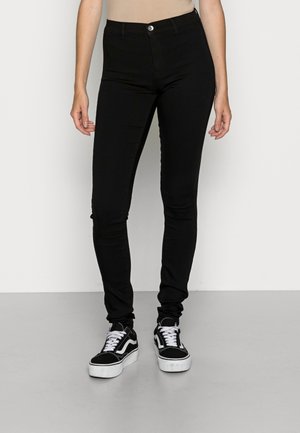 Personne portant un pantalon noir slim et des baskets noires et blanches, debout devant un fond clair uni.