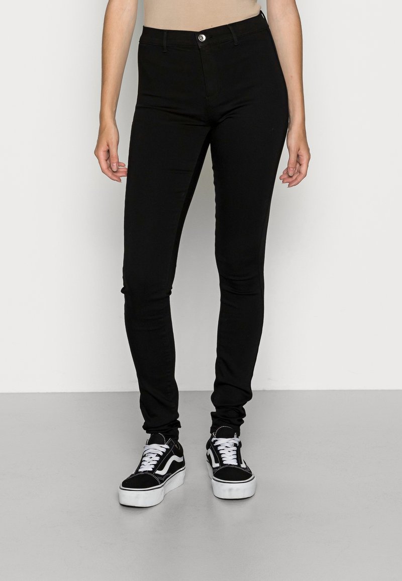 Personne portant un pantalon noir slim et des baskets noires et blanches, debout devant un fond clair uni.