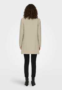 Beżowy długi płaszcz o dopasowanym fasonie, z długimi rękawami i prostym krojem, zestawiony z czarnymi spodniami i botkami za kostkę.
