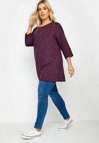 Långärmad bordeauxtröja med silverprickar, kombinerad med slim fit blåjeans och vita sneakers. Enkelt bakgrund.