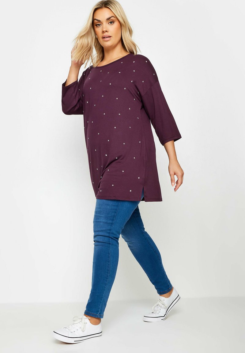 Långärmad bordeauxtröja med silverprickar, kombinerad med slim fit blåjeans och vita sneakers. Enkelt bakgrund.