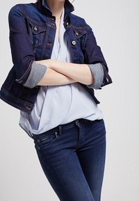 Giacca di jeans blu scuro con accessori dorati, maniche arrotolate e due tasche frontali. Indossata sopra una blusa azzurro chiaro e jeans blu scuro.