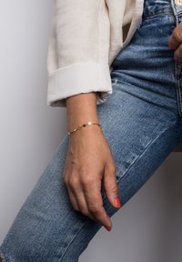 Kuldne käevõru ringikujuliste linkidega, kantud randmel. Raheline kangas särk üles keritud varruka ja sinised denim teksad on kaadris nähtavad.