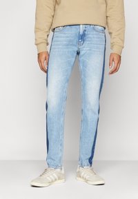 Ljust blå denimjeans med rak passform, som har mörkare sidpaneler, standardfickor och dragkedja, i kombination med beige sneakers.