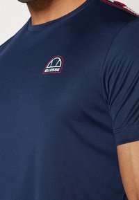 Námořnická modrá sportovní tričko vyrobené z hladkého materiálu. Obsahuje malé logo Ellesse v bílá a červené barvě, s výrazným detailem na ramenou.