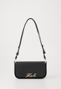 Black leather handbag with a flap design, featuring a gold signature logo. Adjustable strap and smooth texture. Minimalist aesthetic.