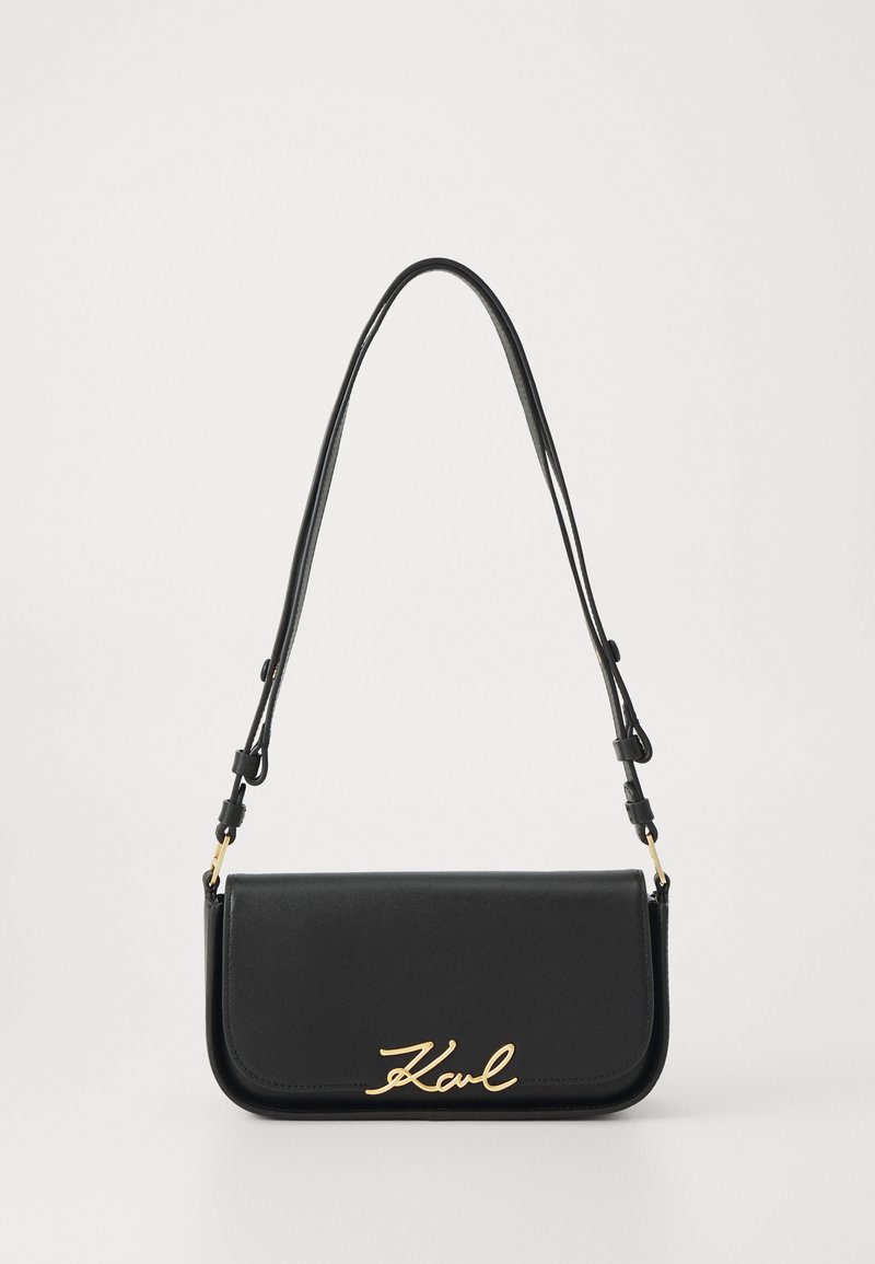 Black leather handbag with a flap design, featuring a gold signature logo. Adjustable strap and smooth texture. Minimalist aesthetic.