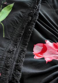 Zwarte stof met franjes, versierd met levendige bloemenpatronen in tinten roze en groen, waardoor een textuur en gelaagd uiterlijk ontstaat.