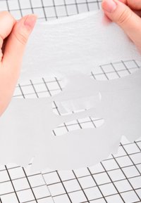 Maschera trasparente per il viso tenuta tra le dita, con ritagli per occhi, naso e bocca. Texture liscia, con una leggera lucentezza, su uno sfondo a griglia.