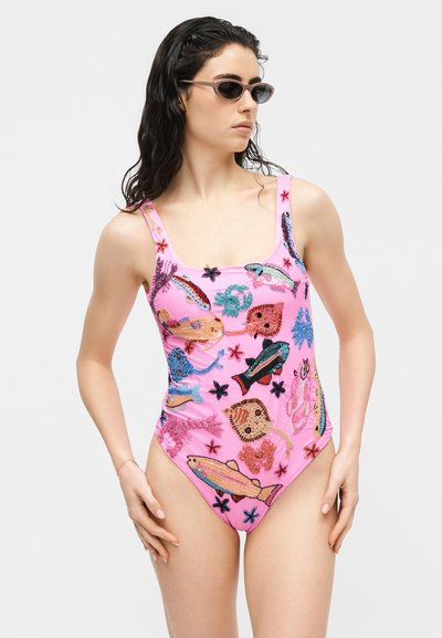 Γυναίκα με ροζ μαγιό στολισμένο με πολύχρωμα κεντημένα ψάρια και θαλάσσια πλάσματα, ποζάρει φορώντας γυαλιά ηλίου μπροστά σε απλό φόντο.