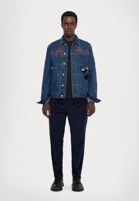 Denim jacket with red floral embroidery, two front pockets, and a slightly loose fit, paired with navy trousers and black shoes.