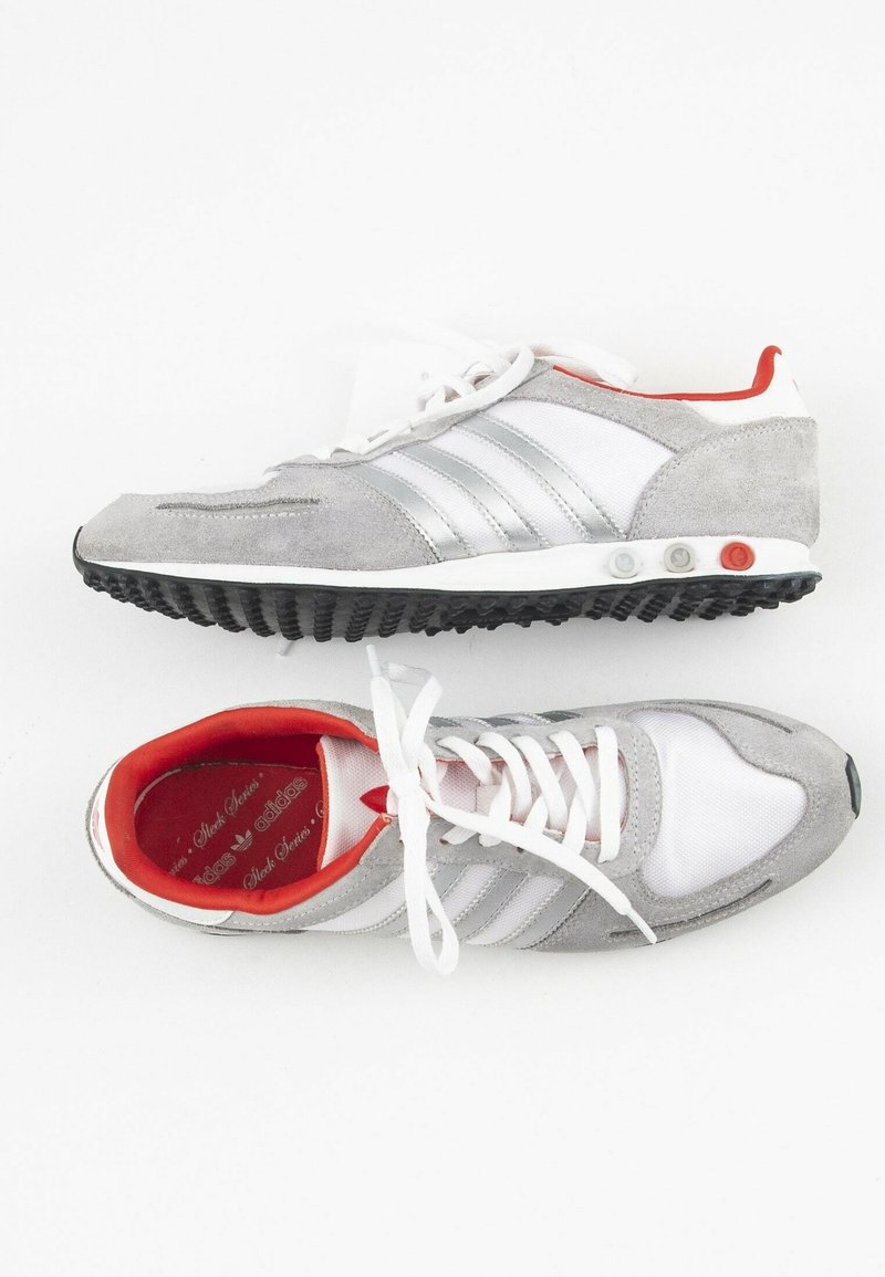 Šedý semišový a bílý síťovaný teniska se stříbrnými pruhy, červenou podšívkou a strukturovanou černou gumovou podrážkou. Má tkaničky a polstrovaný límec.