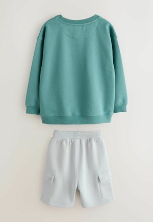 Türkisfarbener Pullover mit rundem Ausschnitt und langen Ärmeln, kombiniert mit hellgrauen Shorts mit Seitentaschen und elastischem Bund.