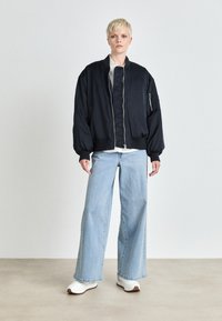 Giacca bomber blu navy con zip frontale, polsini a costine e fodera imbottita, abbinata a jeans a gamba larga blu chiaro e sneakers bianche.