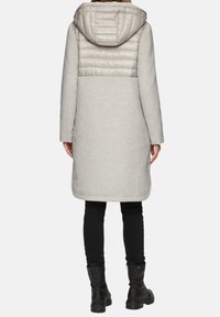 Lichtgrijze jas met een gevoerde capuchon en bovenrug. Heeft een getailleerde snit, lange mouwen en zijzakken. Gekocht over zwarte leggings en laarzen.