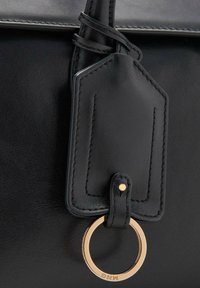 Detalle de bolso de cuero negro que presenta una etiqueta rectangular para equipaje con costuras y un anillo de metal dorado, creando un contraste elegante.
