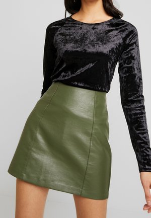 Frau trägt ein schwarzes Samtoberteil mit langen Ärmeln und einen hoch taillierten grünen Leder-Minirock vor einem einfarbigen hellen Hintergrund.